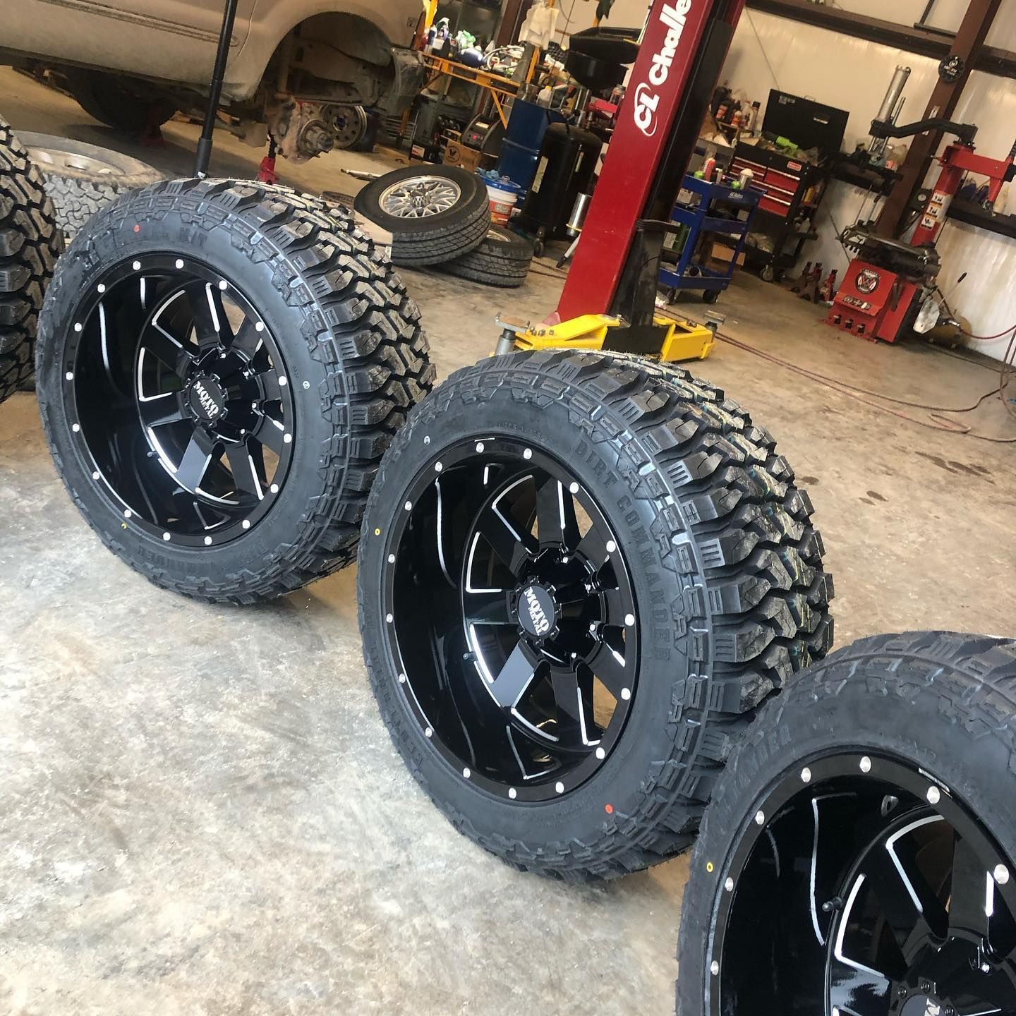 Four black truck wheels with mud tires in a garage.