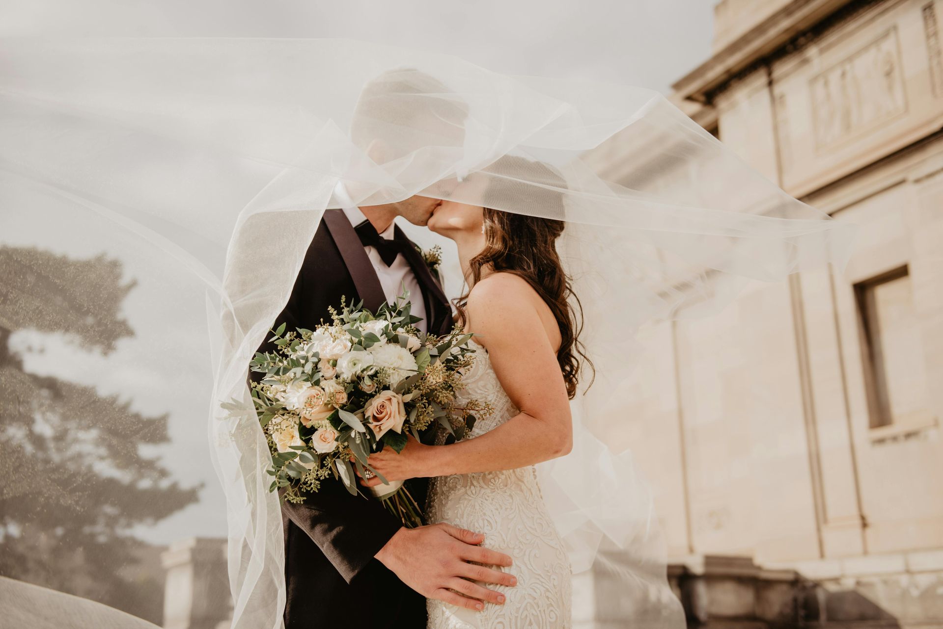 A bride and groom kissing under a veil in front of a building.