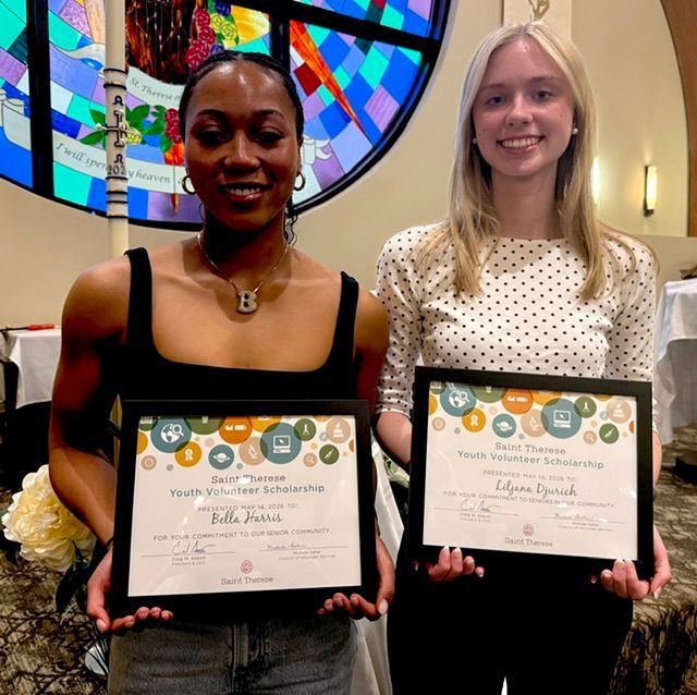 Two individuals stand indoors holding award certificates, smiling at the camera in front of a bookshelf and fireplace.