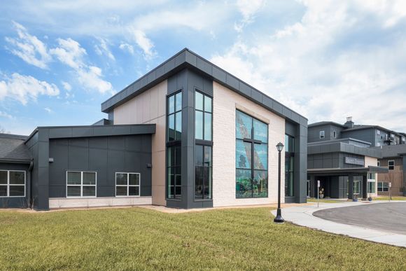 Modern, two-story building with gray metal panels, light stone walls, and large windows under a partly cloudy sky.