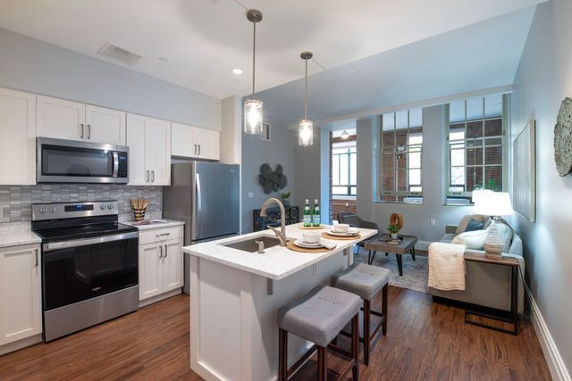 A modern open-plan kitchen and living area with white cabinets, stainless appliances, a kitchen island, and gray sofas.