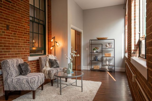 A sunlit seating area with two patterned armchairs, a glass coffee table, a rug, and shelving against a brick wall.