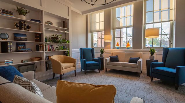 A cozy sitting area with a sofa, two blue armchairs, and a yellow chair arranged on a rug in front of bookshelves.