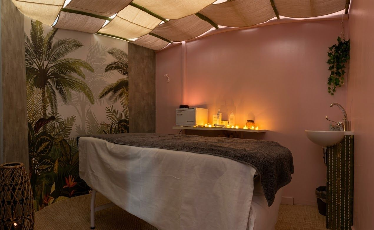 Una sala per trattamenti spa con lettino per massaggi, carta da parati a tema tropicale, luci soffuse e calde e un piccolo lavandino.