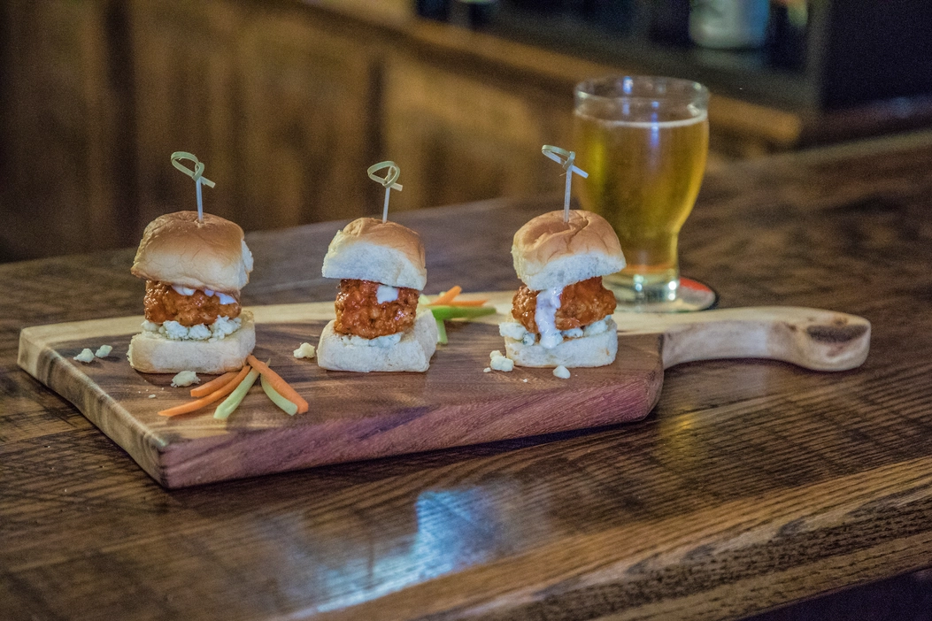 Three sliders are sitting on a wooden cutting board next to a glass of beer.