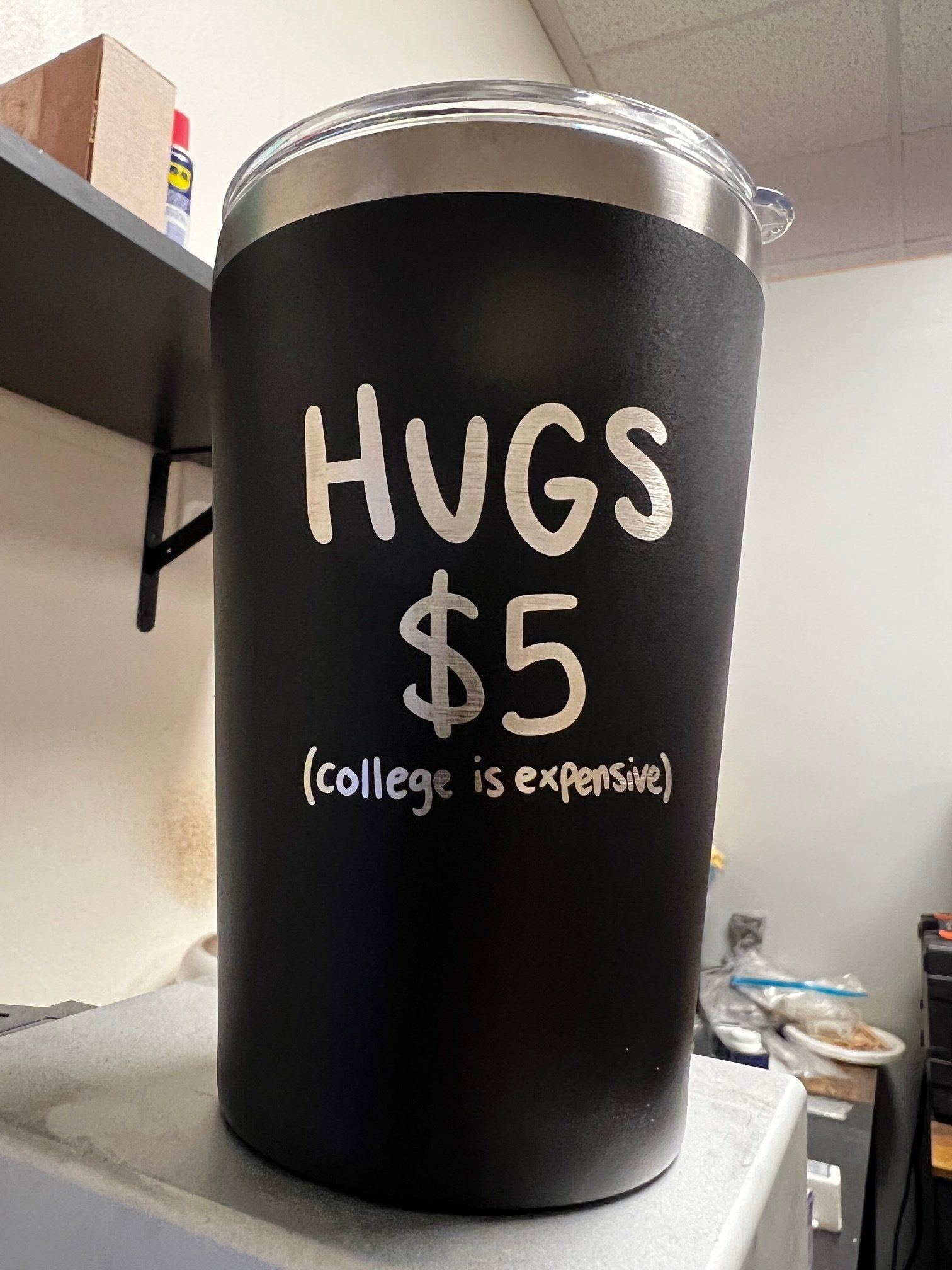 A black tumbler that says `` hugs $ 5 ( college is expensive ) '' is sitting on a counter.