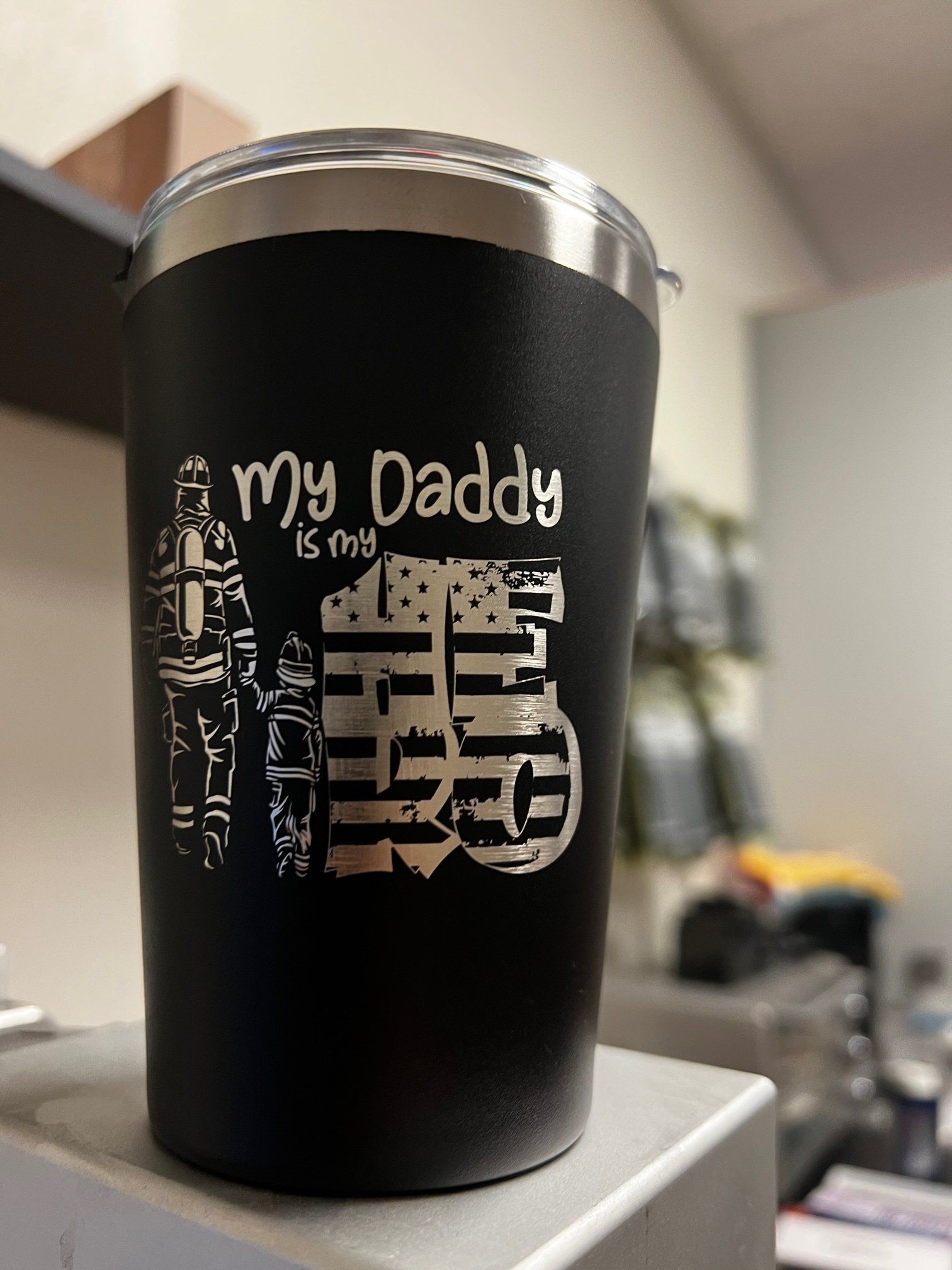 A black tumbler with the words `` my daddy is my hero '' on it is sitting on top of a table.