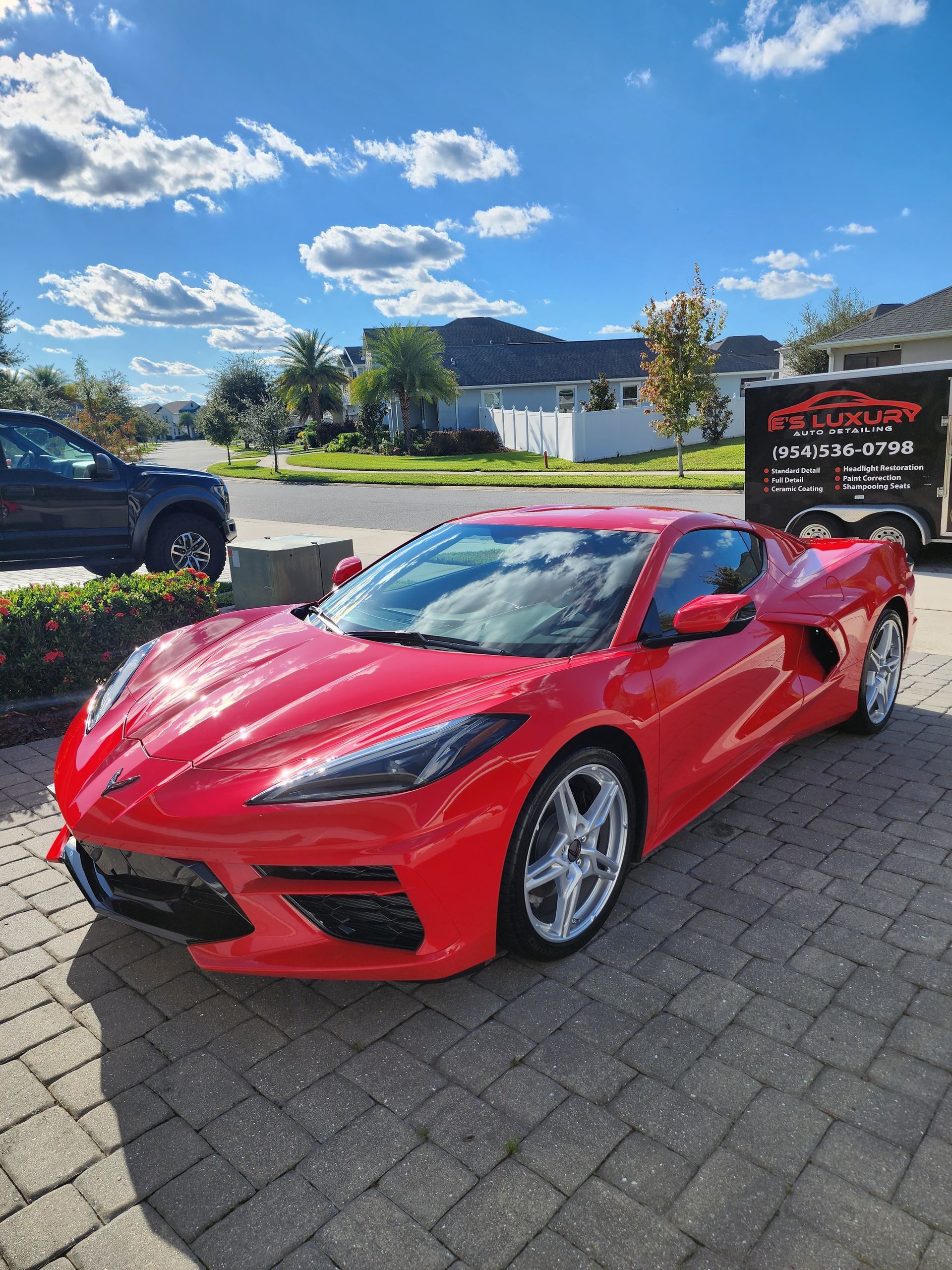 Red Corvette sports car parked on a brick driveway under a blue sky, with a black trailer in the background.