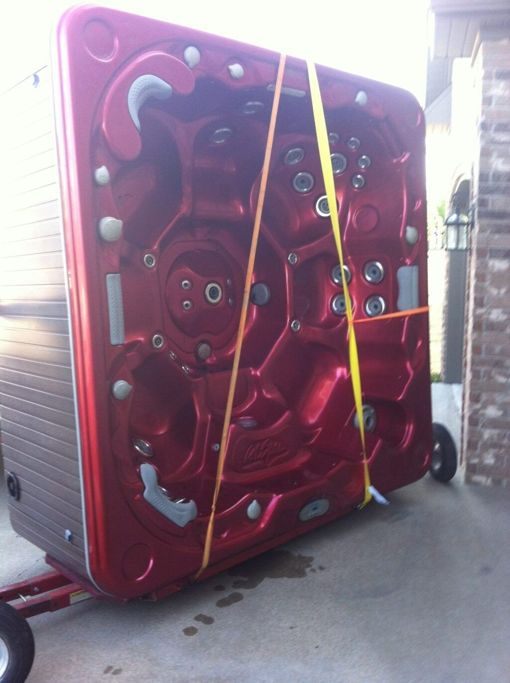 A bright red hot tub secured with yellow straps to a trailer on a concrete patio next to a brick wall.