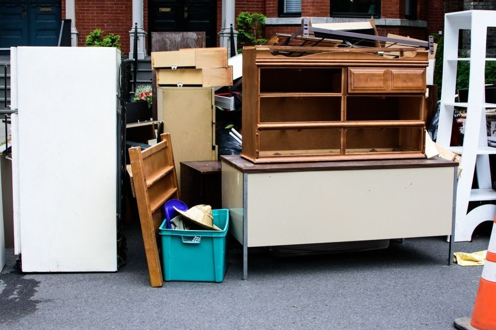 A sidewalk pile of discarded items, including a refrigerator, wooden furniture, a teal plastic bin, and a white shelf.