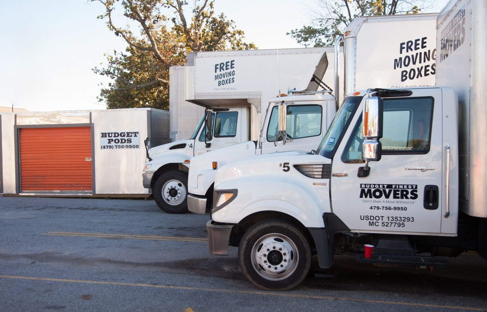 A line of white moving trucks and a storage unit in a parking lot, with text advertising free boxes on the truck exteriors.