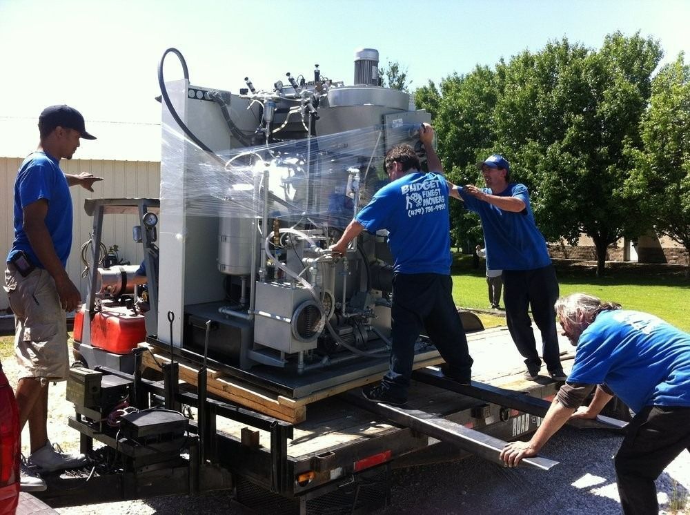 Four people in blue shirts move a large, shrink-wrapped piece of machinery onto a utility trailer outdoors.