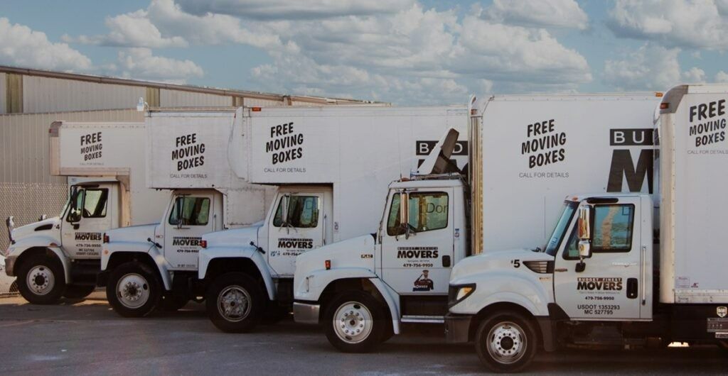A row of five white moving trucks parked in a line against the side of a building under a cloudy sky.