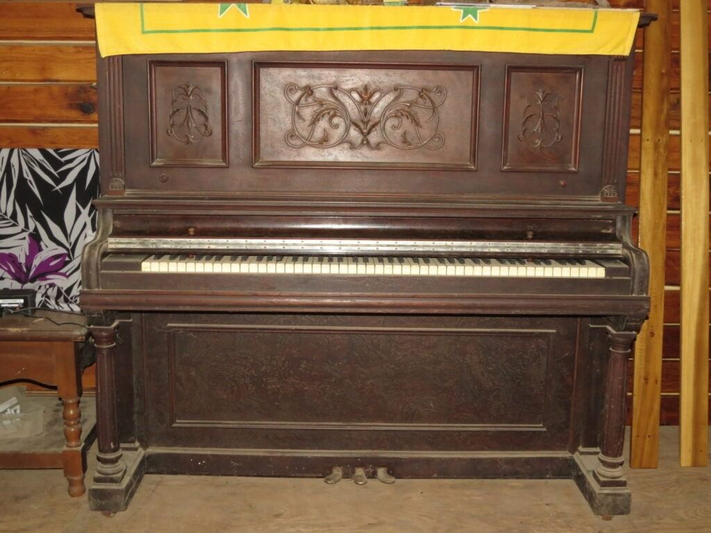 An old, dark wood upright piano with decorative carvings stands against a wooden wall, topped with a yellow cloth.
