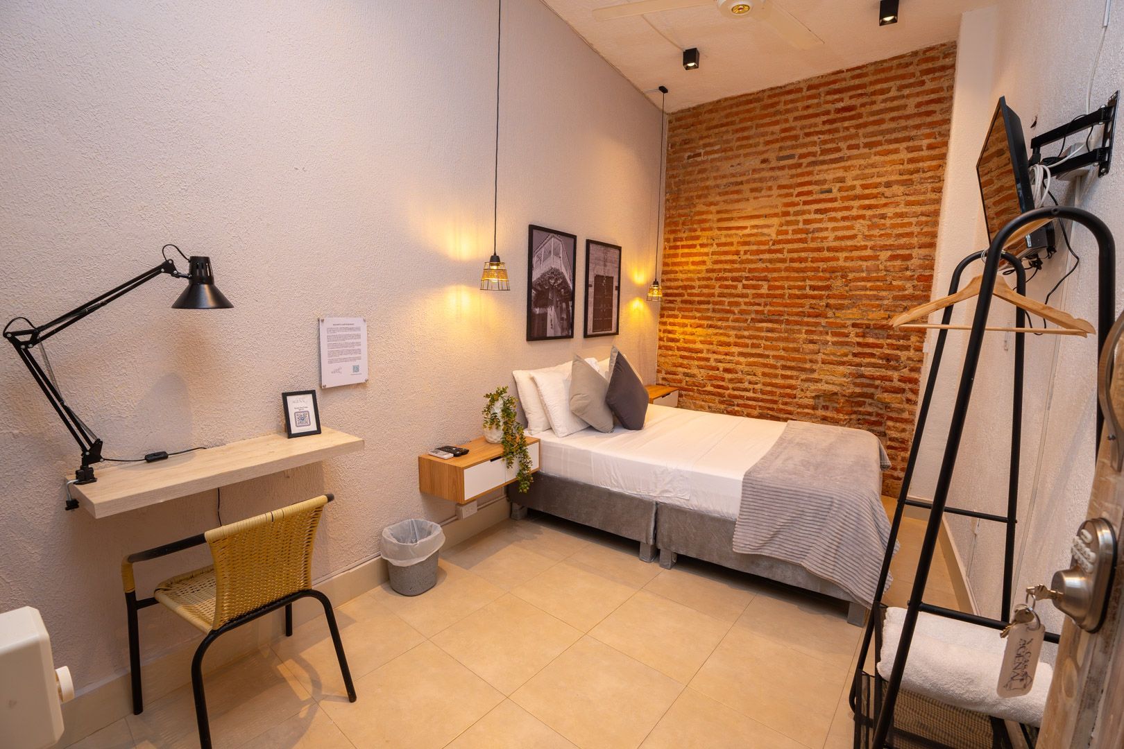 Small hotel room with bed against brick wall, desk, chair, and clothes rack.