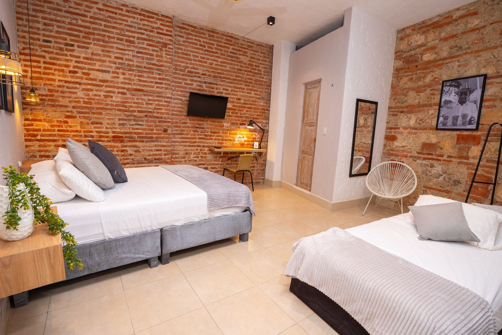 Hotel room with two beds, brick walls, desk, TV, and decorative plants.