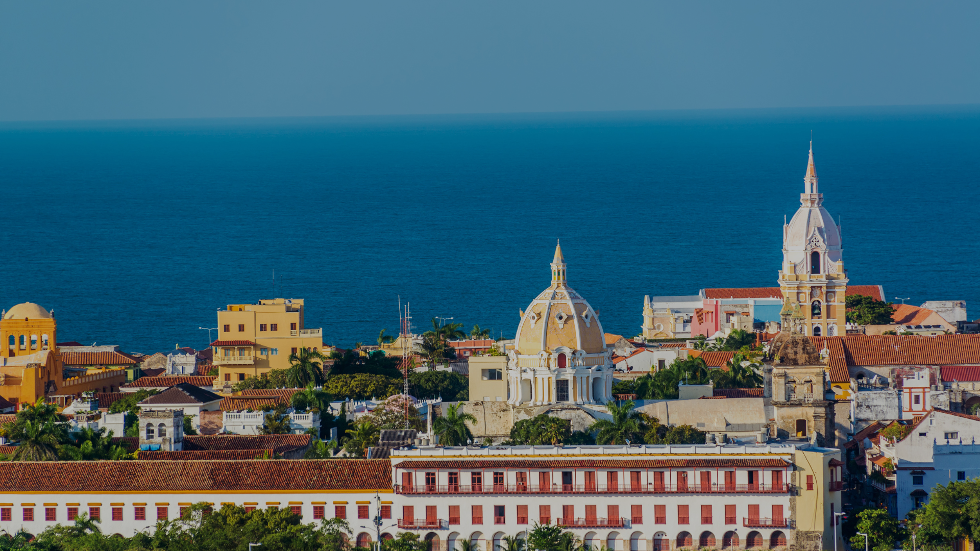 City skyline with buildings, domes, and a church tower against the ocean.