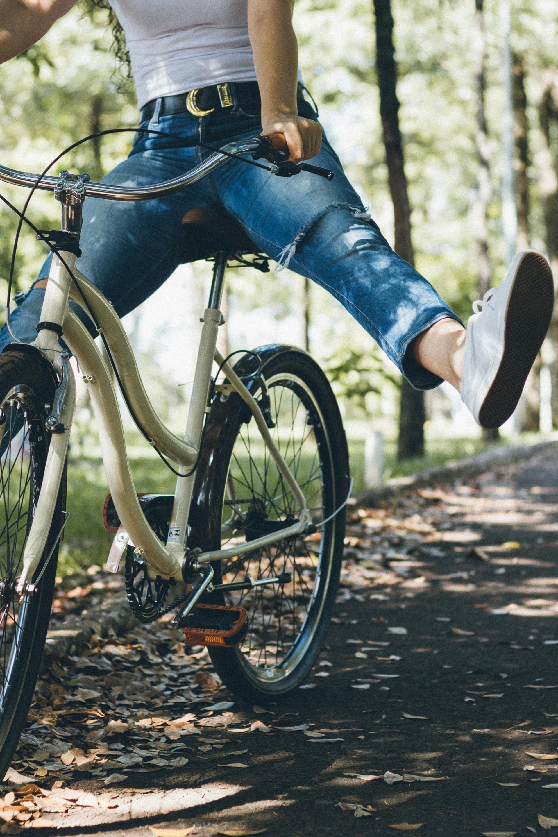 Person riding a bicycle, one leg extended. Wearing jeans, white top, and sneakers. Outdoor setting with trees.