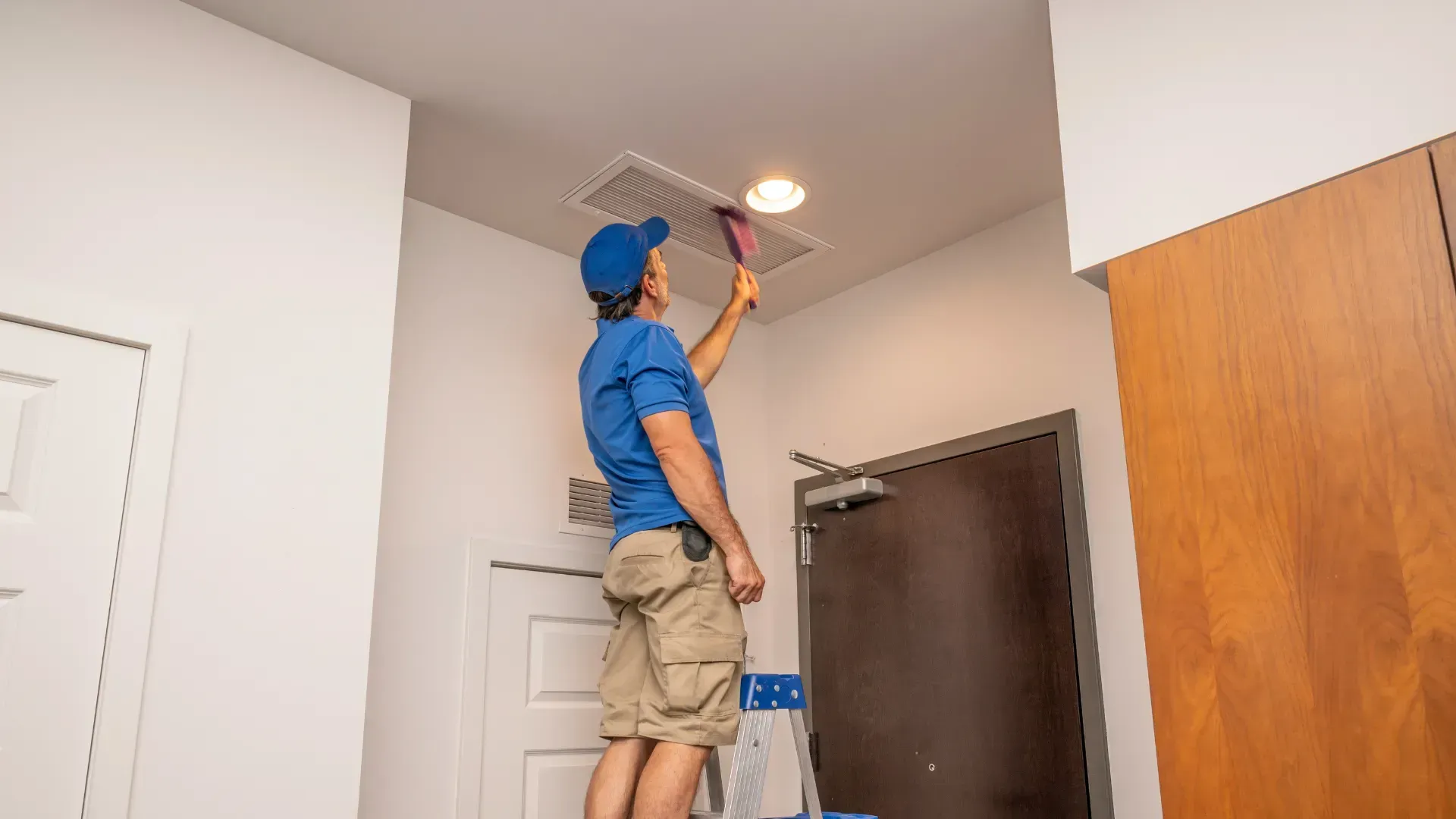 A man is cleaning the air vent on the ceiling of a room.