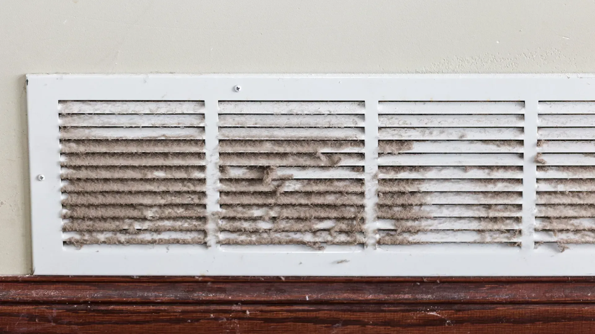 A dirty air vent on a wall next to a wooden trim.