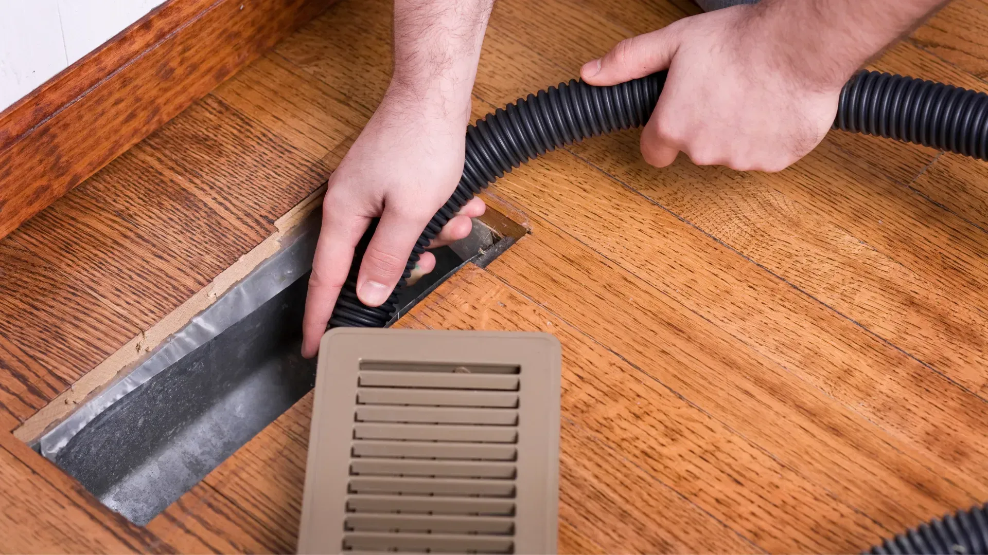 A person is using a vacuum cleaner to clean a vent on the floor.