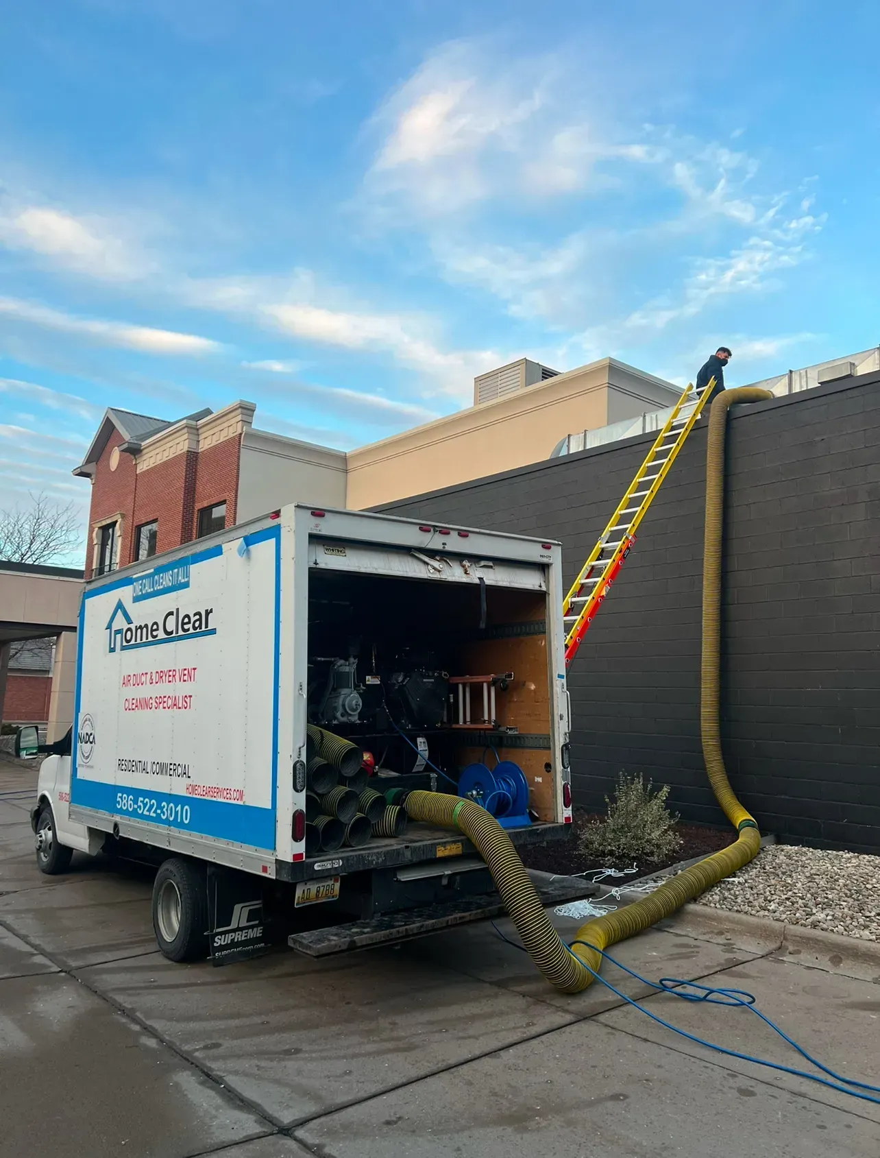 A white Home Clean truck with a yellow hose connected to a wall; a person on a ladder is at the top.