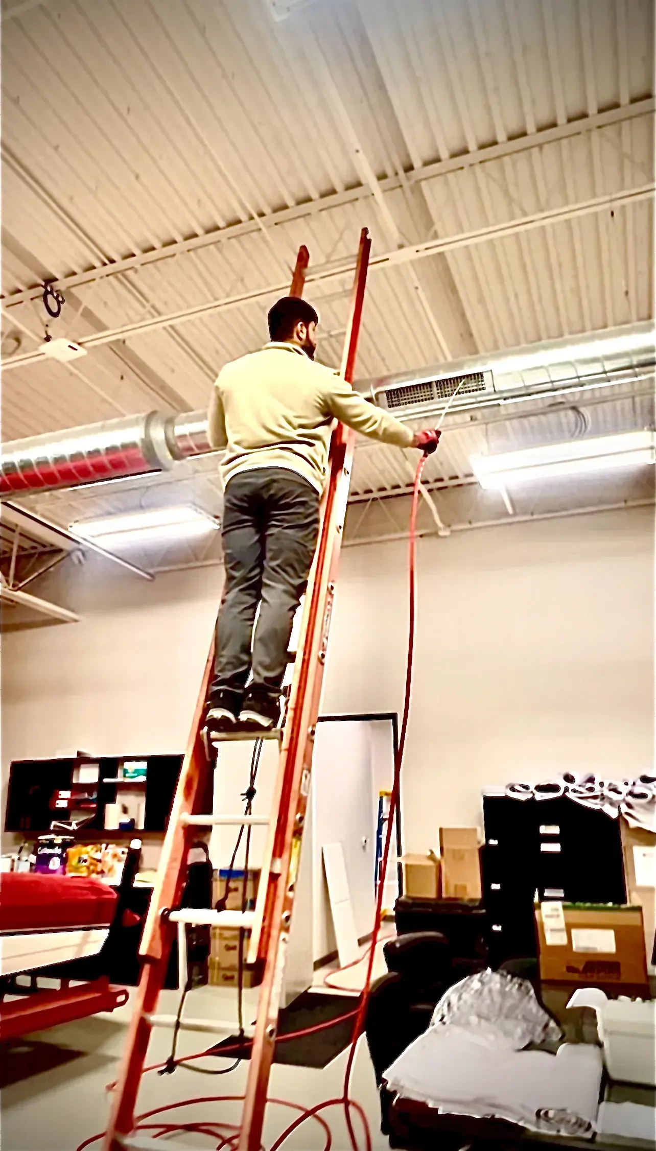 Man on orange ladder reaching for something attached to ceiling in a warehouse.