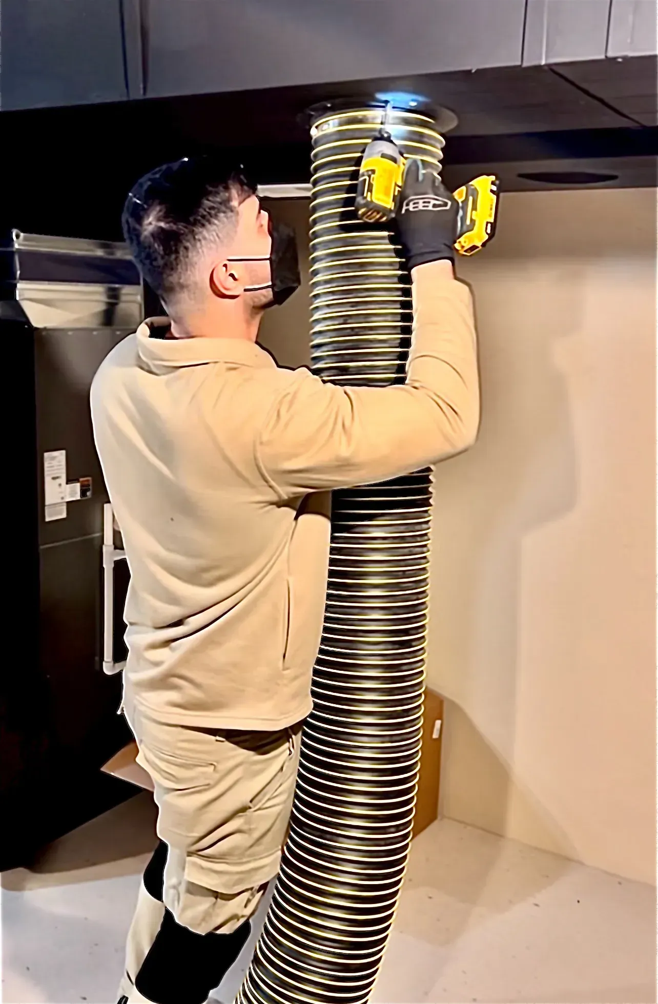 Man in work clothes attaching a flexible metal vent to a ceiling with a drill.