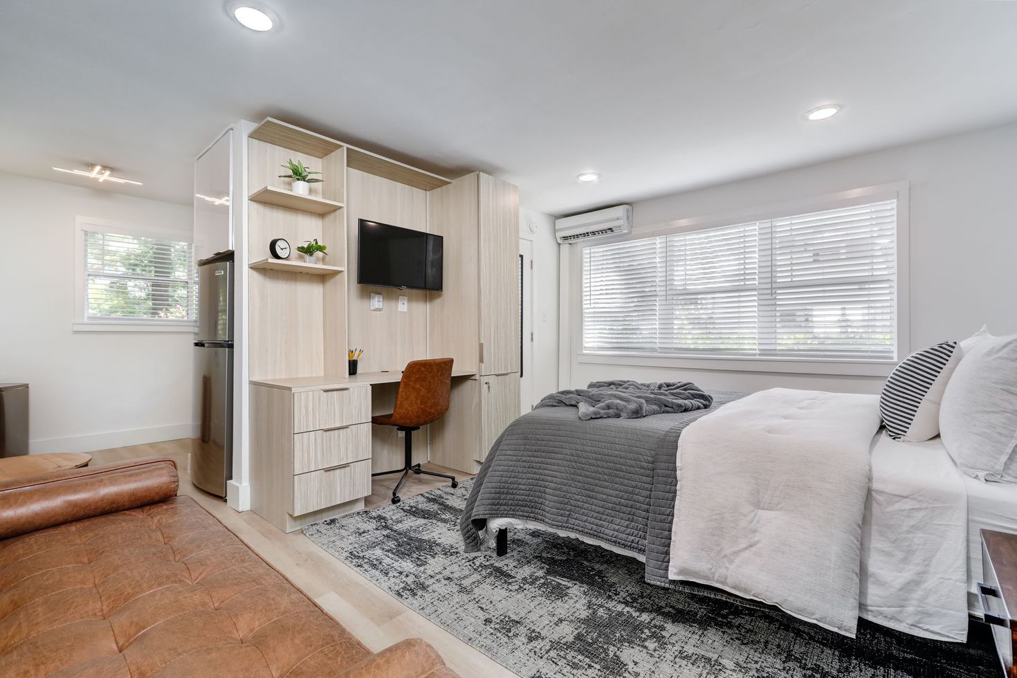A studio apartment with bed, desk, and built-in shelving unit, next to a window with blinds.