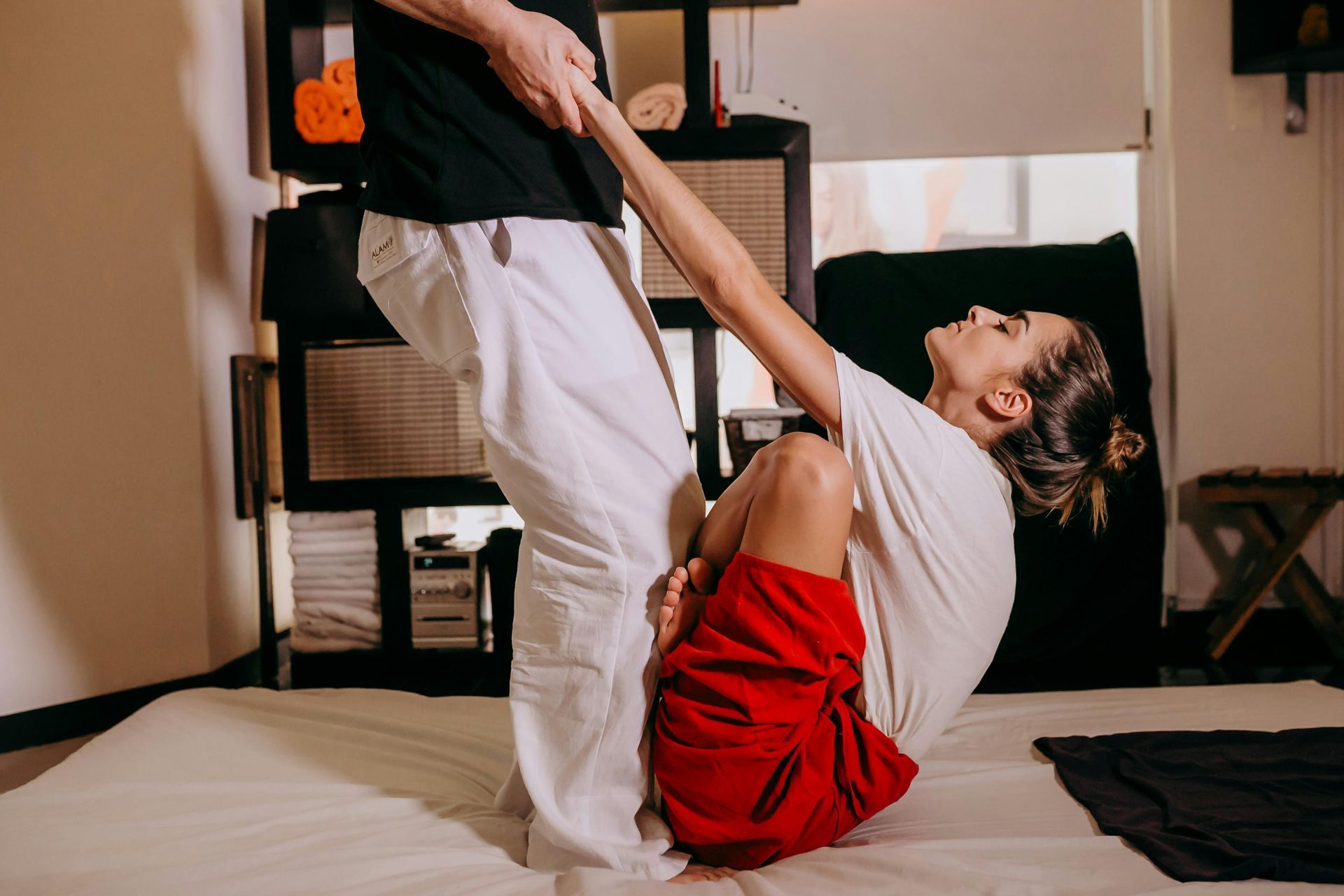 Woman receiving Thai massage: held by practitioner, bent backward on mat, indoors.