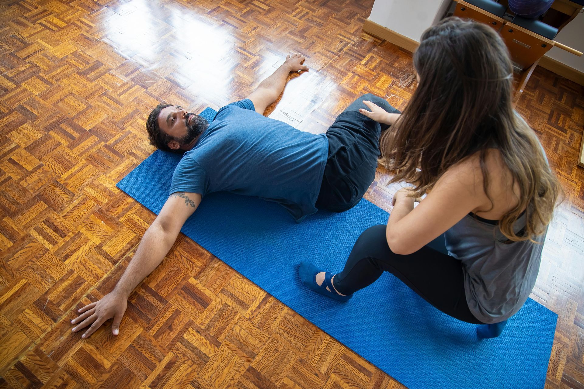 A woman helps a man stretch on a blue yoga mat in a room with wooden floors.