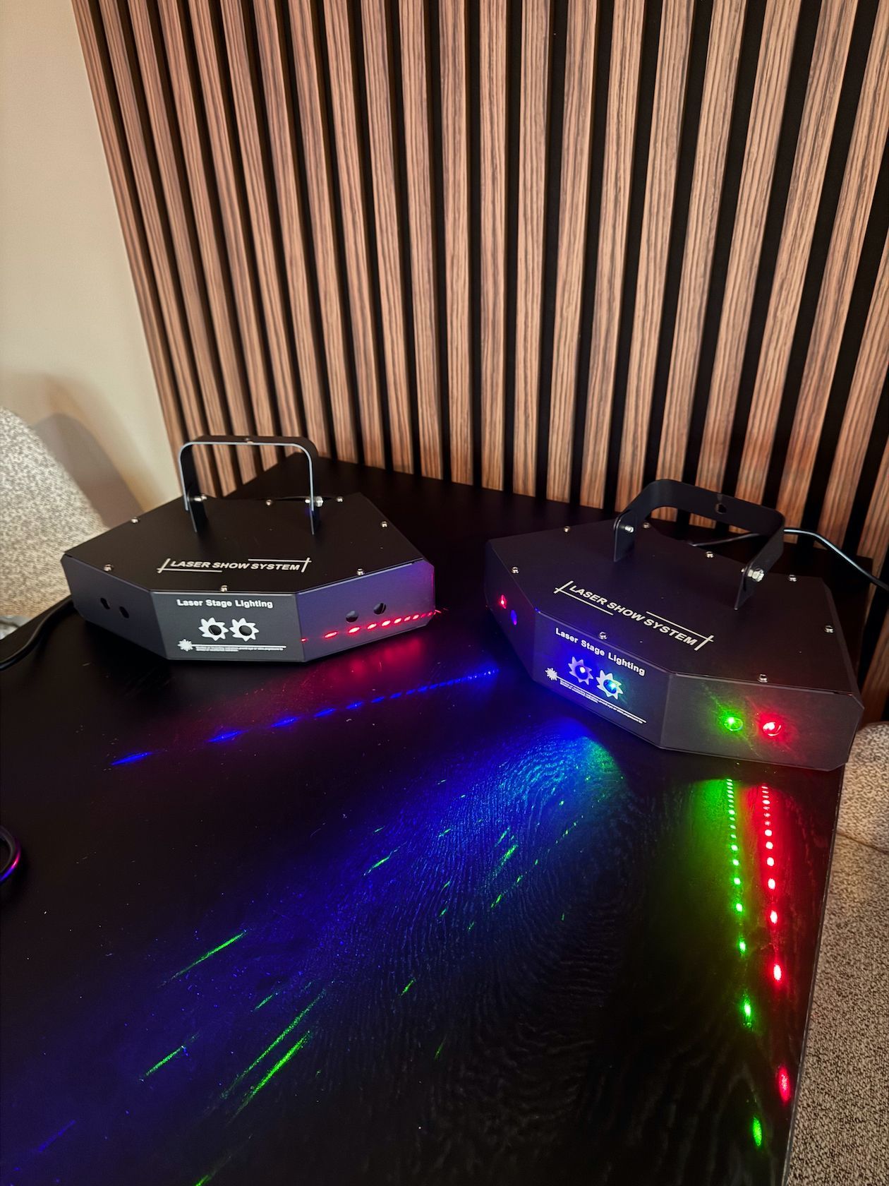 Twee zwarte laserprojectoren die rode, groene en blauwe lichtstralen op een donker oppervlak projecteren.