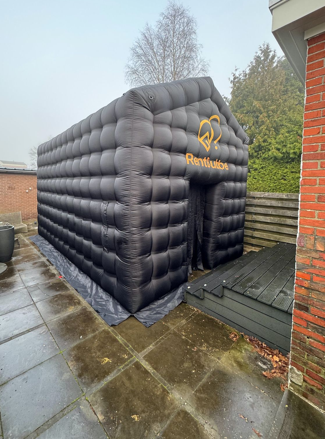 Zwarte opblaasbare kubusvormige constructie op het terras, deuropening zichtbaar. Hartvormig logo aan de zijkant. Natte ondergrond, bakstenen muur.