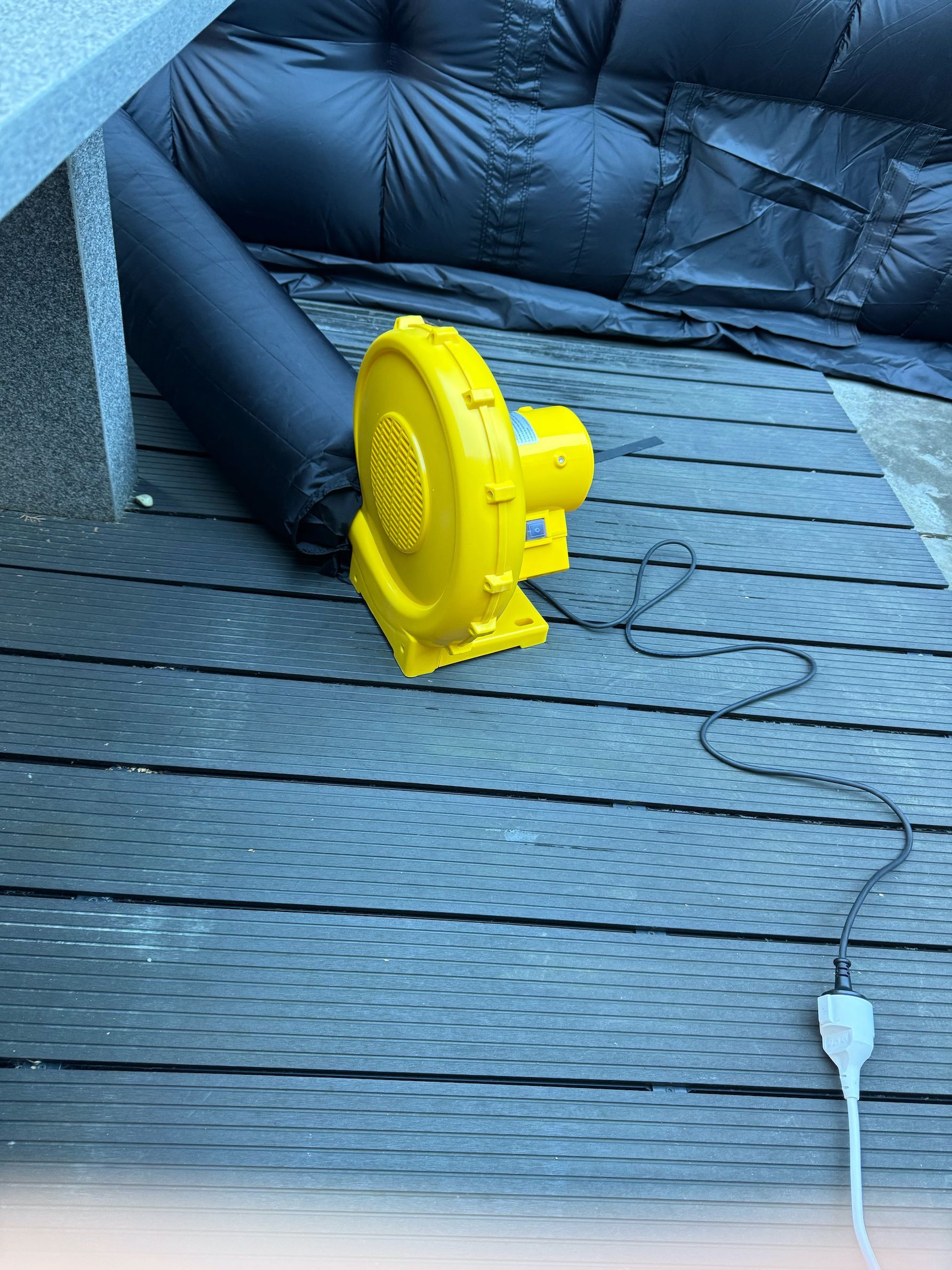 Gele elektrische ventilator aangesloten op een zwarte opblaasbare constructie op een houten terras.