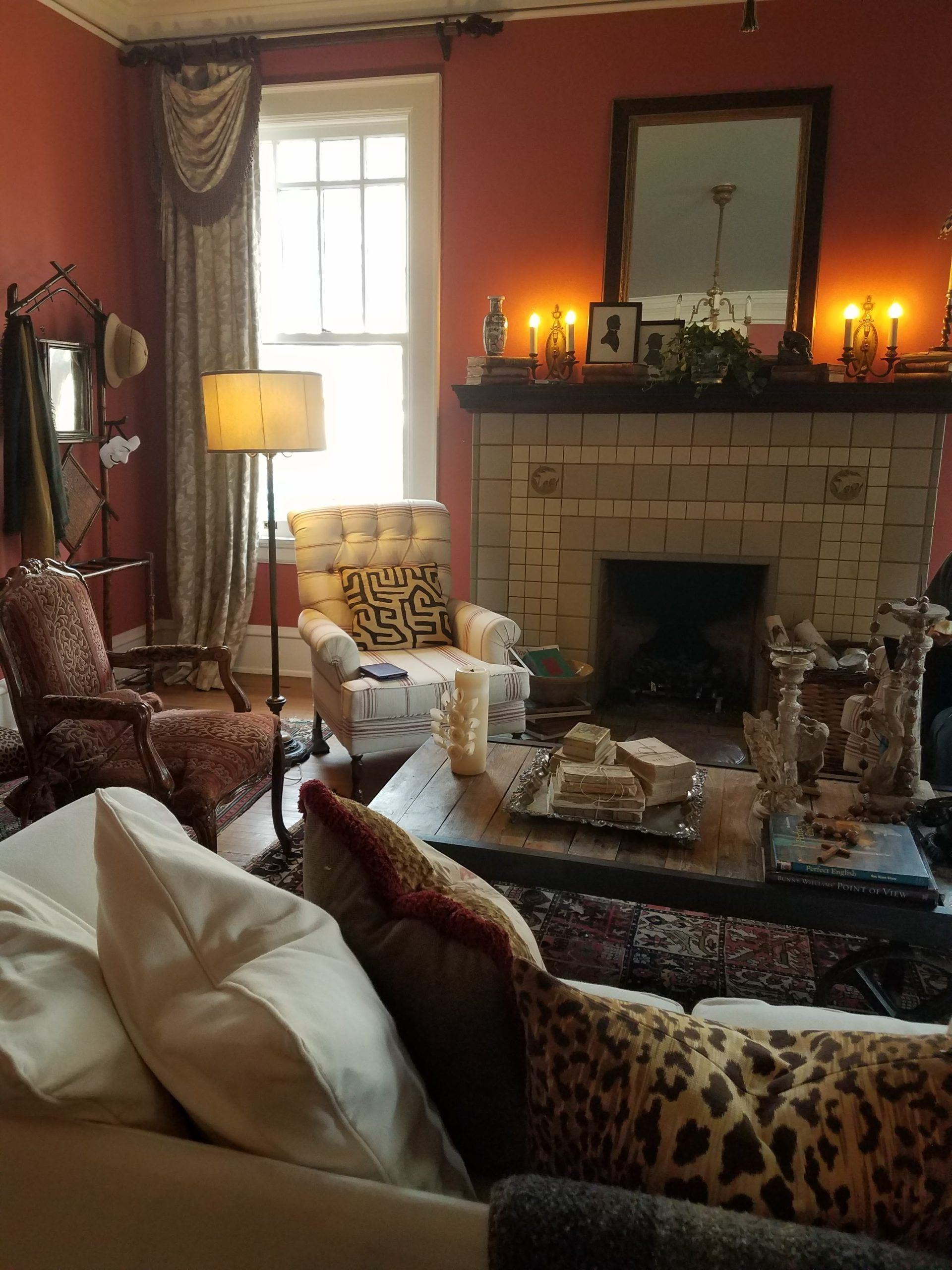 Cozy living room with fireplace, red walls, and patterned furniture. Warm lighting, candles, and window.