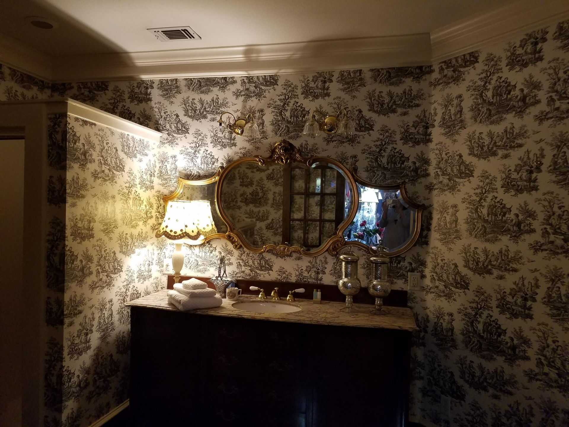 A gilded mirror hangs above a dark wood cabinet, with a lamp and decor against toile wallpaper.