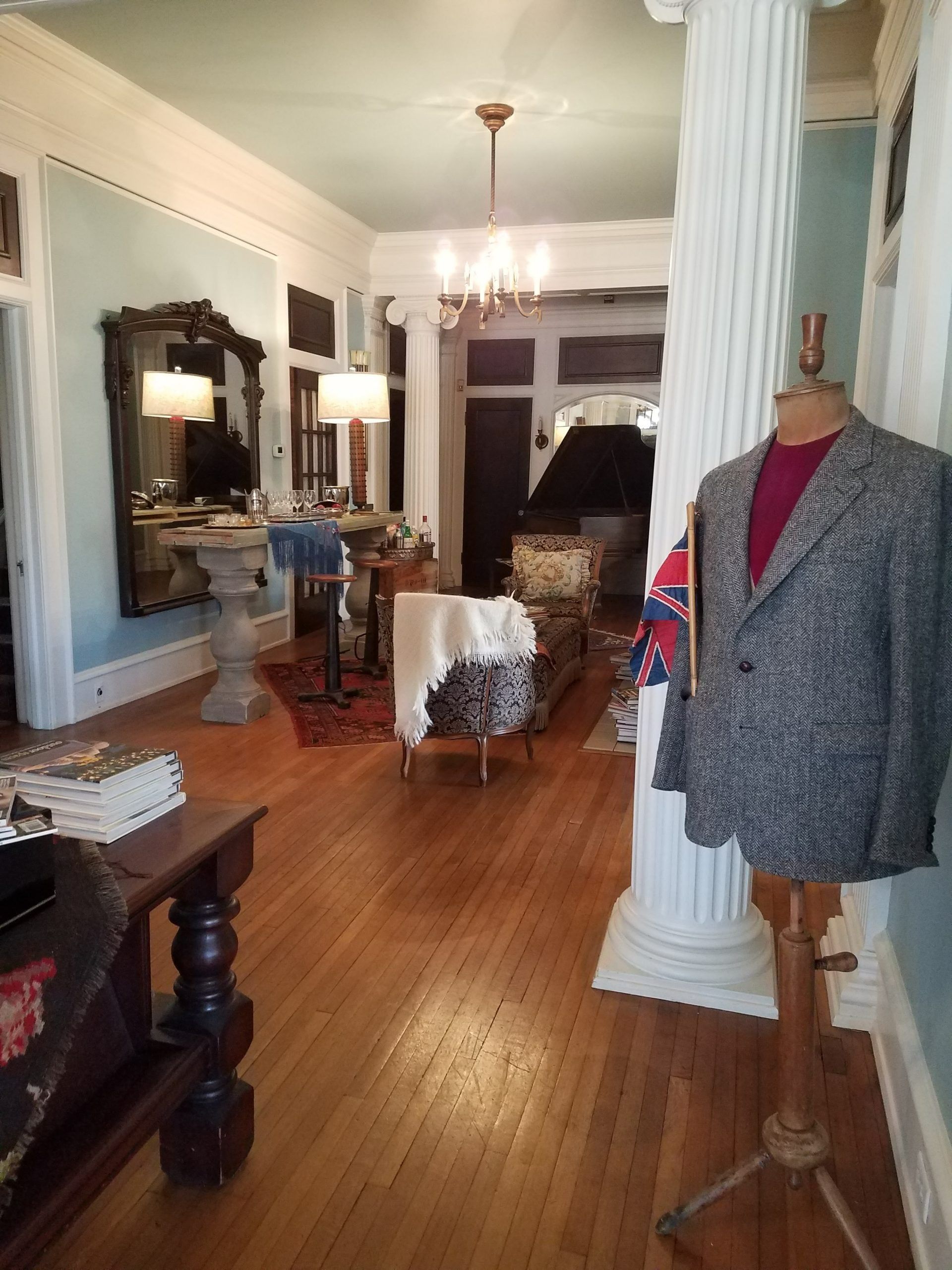 A room with hardwood floors, a vintage mannequin wearing a tweed blazer, and an entryway with columns and ornate furniture.