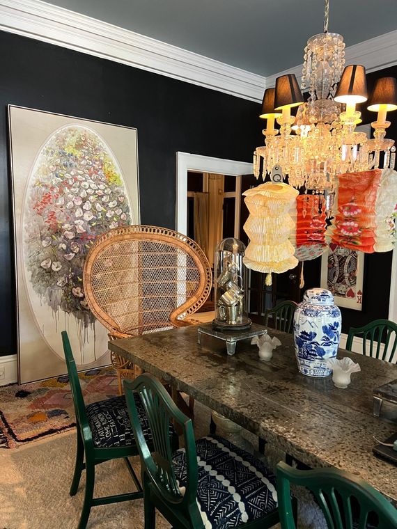 Dining room with dark walls, ornate chandelier, long table, green chairs, and eclectic art.