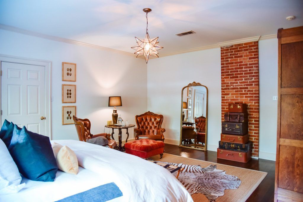 Bedroom with bed, chairs, mirror, and stacked luggage by a brick wall.