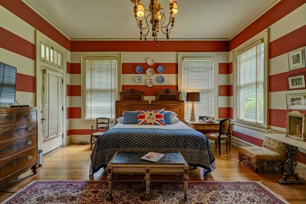 Bedroom with red-striped walls, ornate wooden bed, windows, chandelier, and antique furniture.
