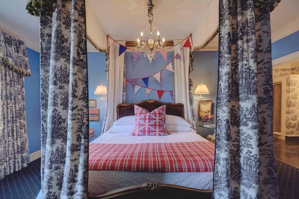 Bedroom with a canopy bed. The bed has red and white linens and a pink patterned pillow. Blue walls and patterned curtains.