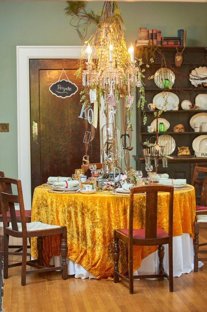 Dining table set with yellow velvet tablecloth, chandelier, and china on shelves.