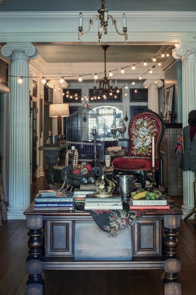Antique table laden with objects, a decorated chair, columns, and string lights in a room.