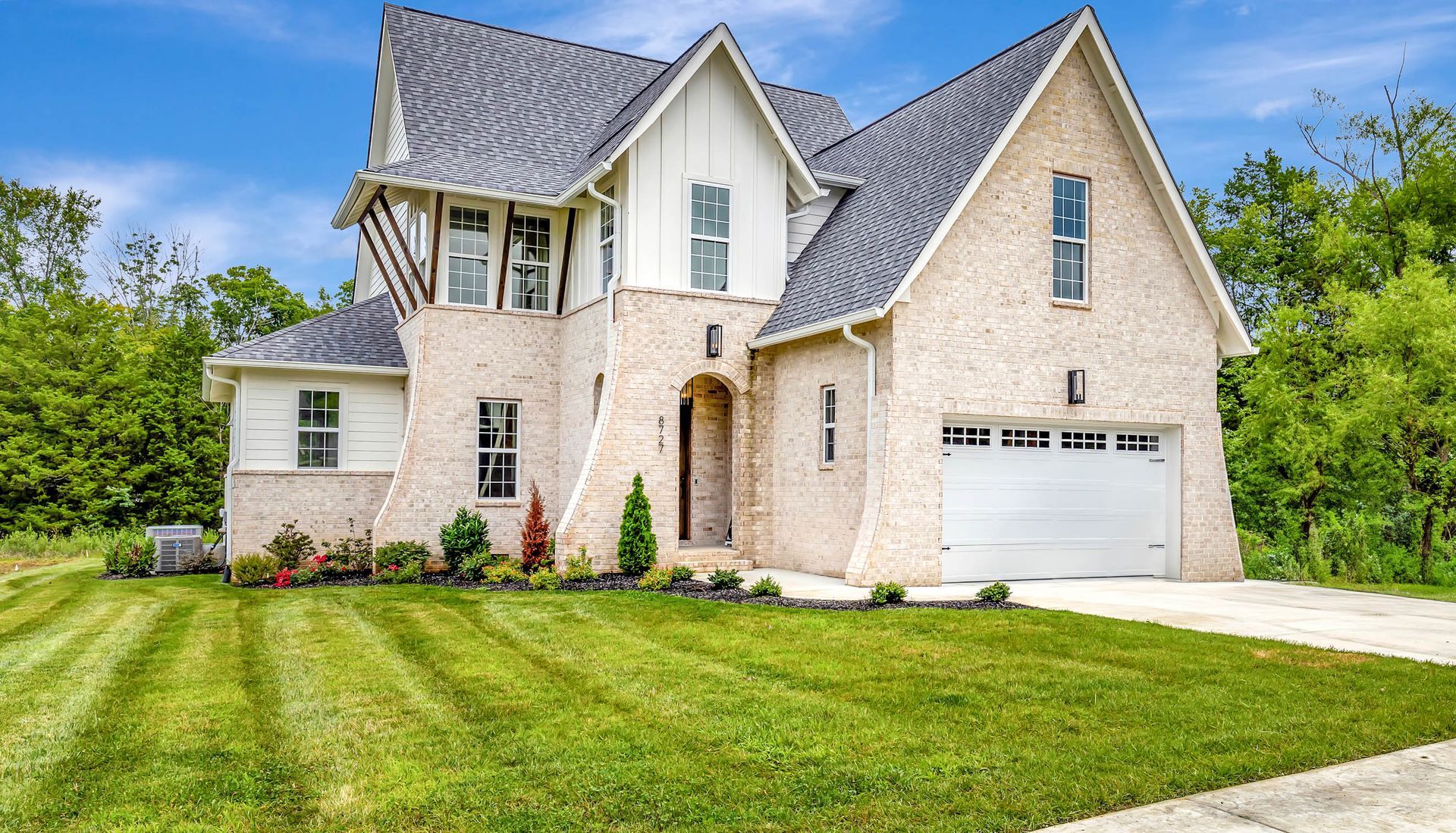 A large brick house with a gray roof and a white garage door is sitting on top of a lush green field.