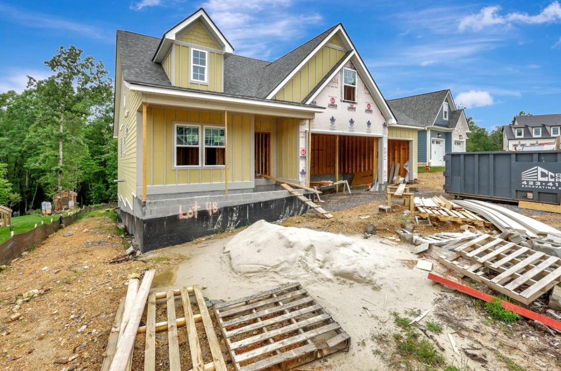 Construction of a light yellow house with attached garage. Pallets and debris are in the foreground.