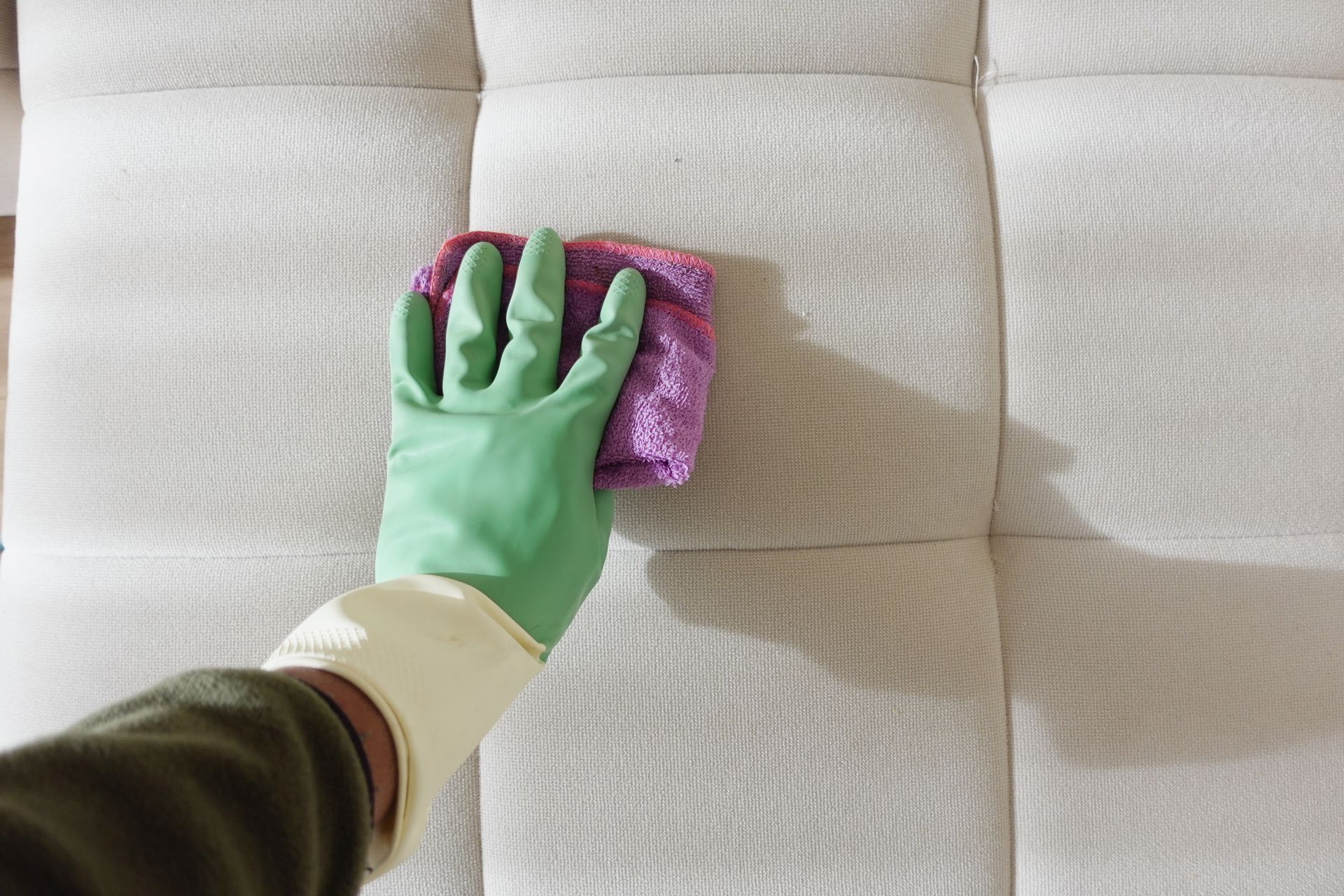 A hand wearing a green glove wipes a sofa with a purple cloth gently. A hand wearing a green glove wipes a sofa with a purple cloth gently.