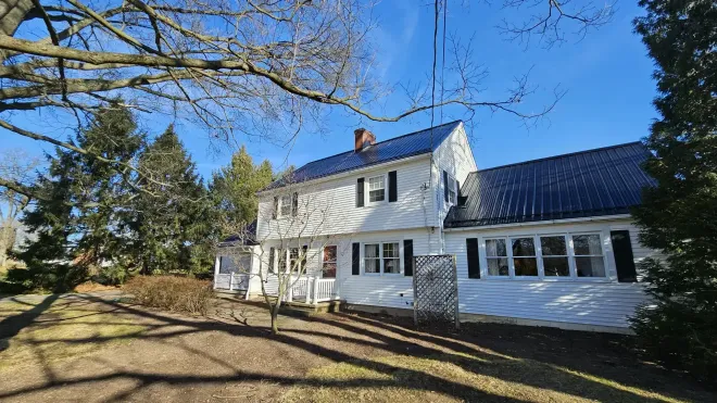 White house with a dark metal roof, set on a sunny property with bare trees and evergreens on a bright, clear day.