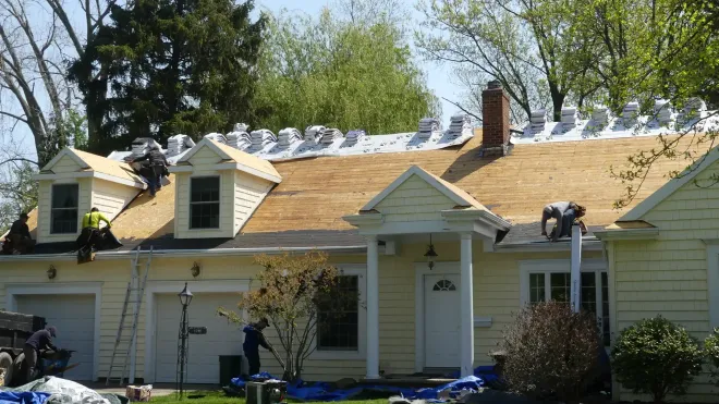 Workers in high-visibility vests perform roof repairs on a yellow house with white trim and dormer windows.