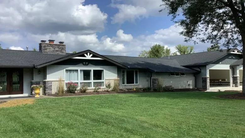 A single-story suburban house with light gray siding, a dark shingled roof, stone accents, and a large lawn under a blue sky.