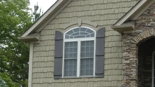 A beige-shingled house exterior featuring an arched window with dark gray shutters and stone masonry on the right.